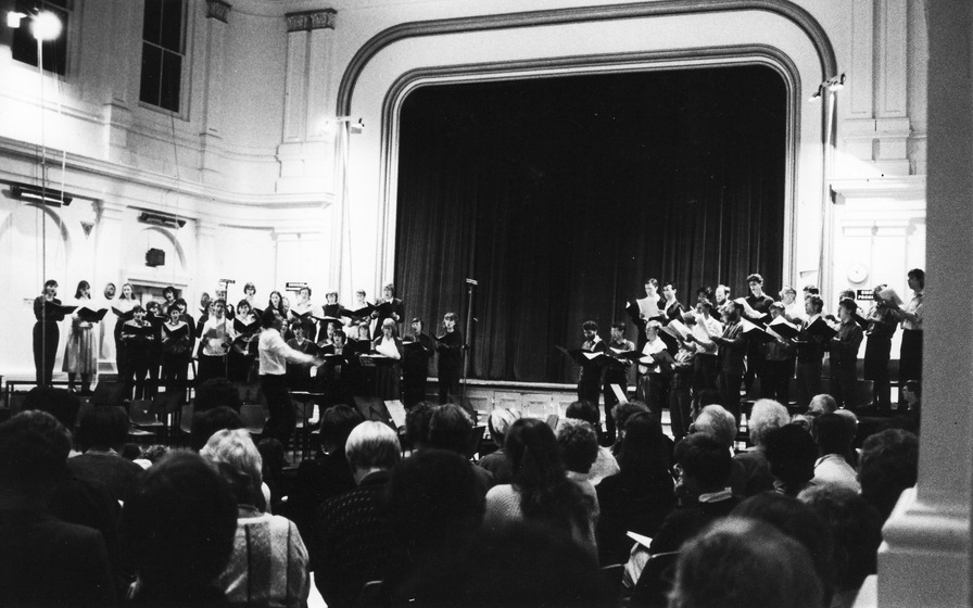 Black and White image of a group of choral singers standing on a stage, split into two groups on the left and right of the room
