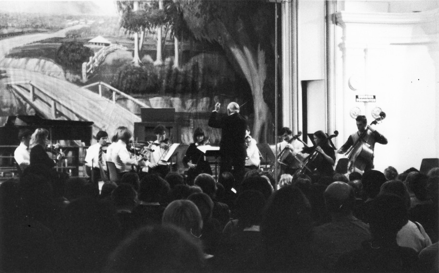 Audience watches a seated string orchestra while a conductor stands at the front with his arm in the air