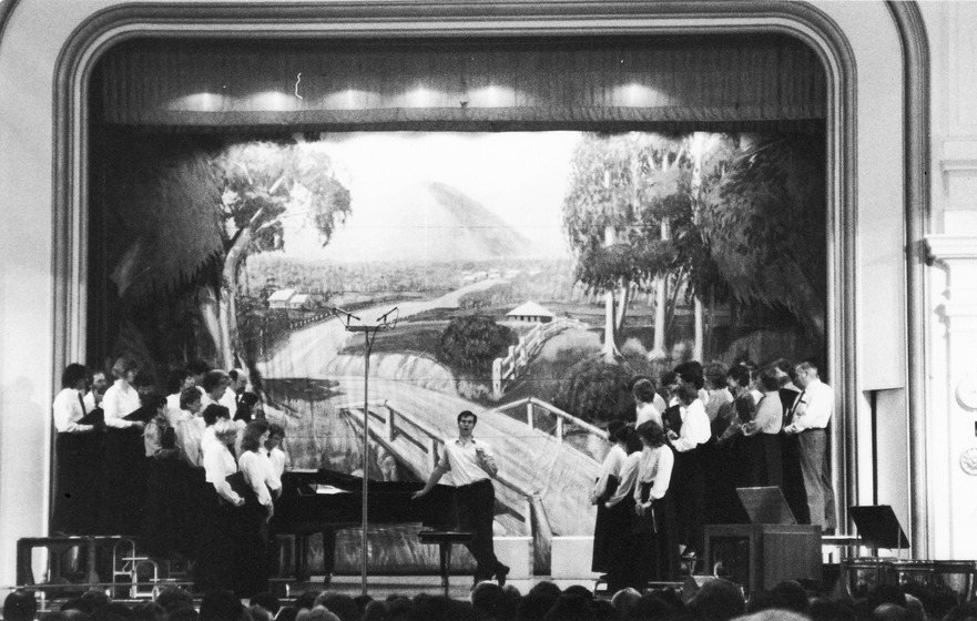 Two groups of singers stand on either side of a stage, looking at a conductor in the middle. A decorative backdrop is featured at the back of the stage