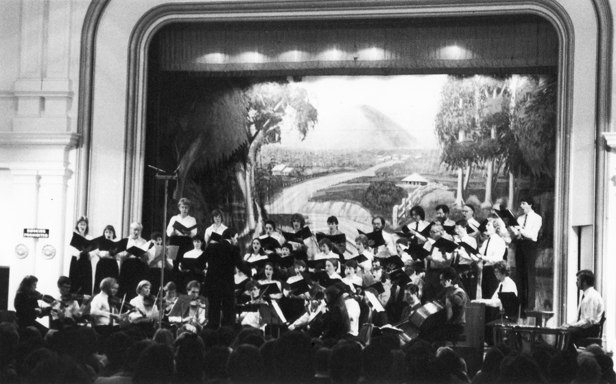 A group of choral singers stand on a stage, led by conductor at the front of the group, with his back to the audience