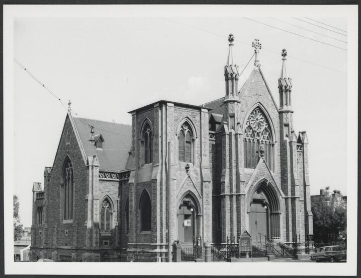 Black and white image of a brick church. The front has a flat facade with a large stain glass window above the arched door.