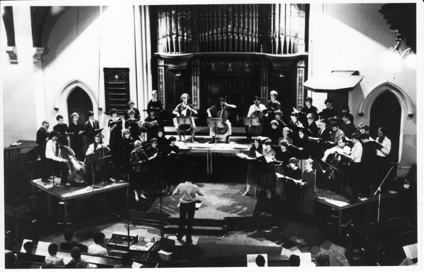 A medium size orchestra sit on a raised platform in front of a large organ at the front of a church. A conductor stands in front of the orchestra, and an audience watches from the pews.