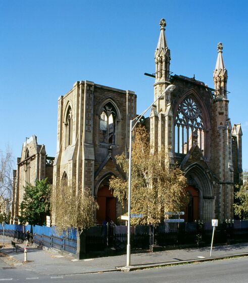 A tall sandstone church stands on a corner block of a street. The church has been gutted by a fire, so only the brick work and wooden doors remain in tact.