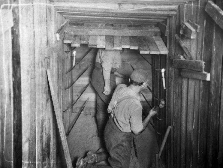 Black and white photograph of two men in a wood panelled tunnel with two candles in brackets, one man hammering.