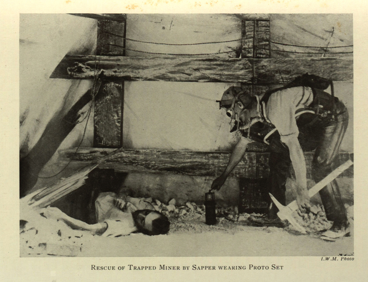 Black and white posed studio photograph of an unconscious man in a tunnel. Another man approaches with mask, pick and pack.