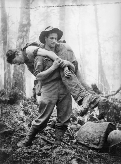 Black and white photograph of a soldier carrying a wounded soldier on his shoulder on a muddy track.