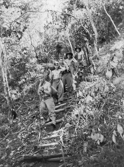 Black and white photograph of six shirtless soldiers carrying supplies down a steep path in the forest.