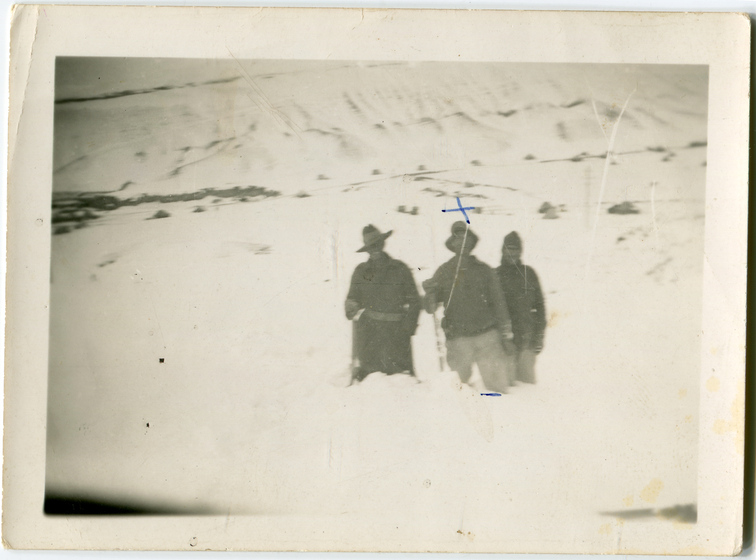 Black and white photograph of three men standing knee deep in snow, one in an Australian army uniform, two holding staffs.