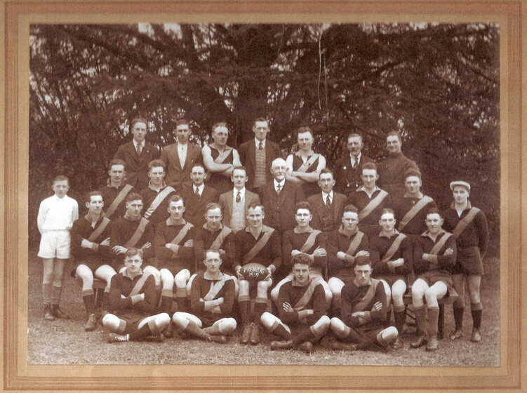 Black and white photograph of a men's football team in guernseys with diagonal stripes. The men are arranged in four rows in a garden.