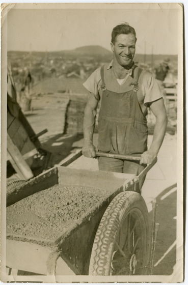 Black and white photograph of a smiling man in overalls pushing a barrow full of wet cement.