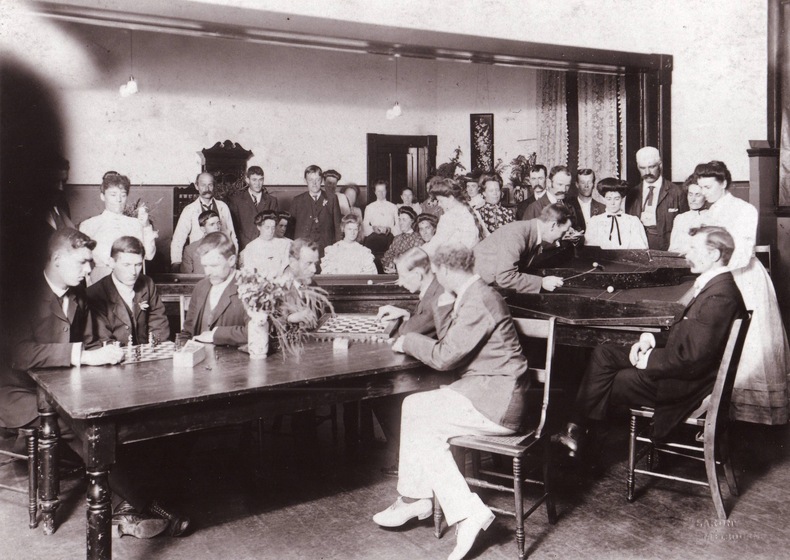 Black and white photograph of a room of men and women playing cards, music and conversing.