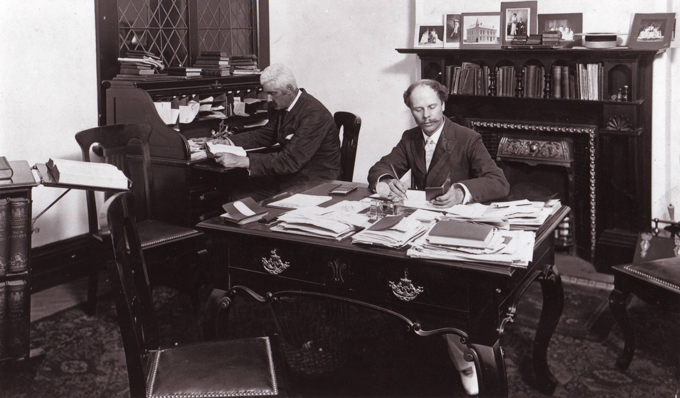 Black and white photograph of two men sitting at desks in a Victorian-style office.