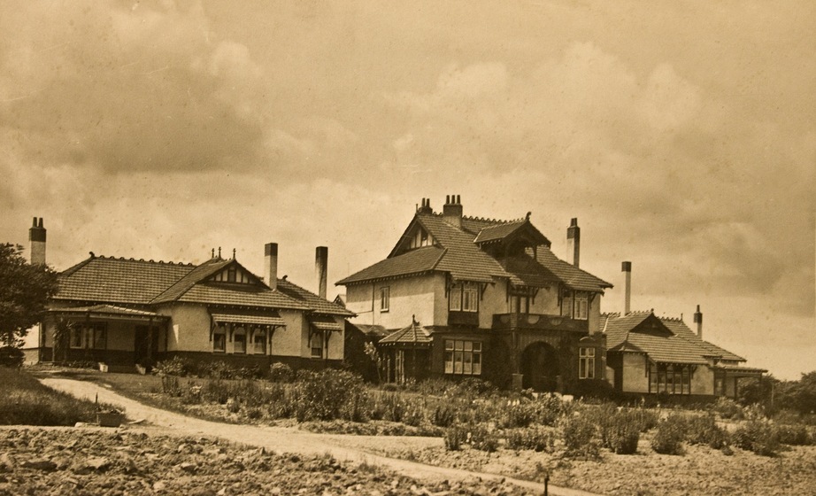 Black and white photograph of three large conjoined 1920s style brick and stucco buildings with extensive terracotta tile rooves in a new garden under a cloudy sky.