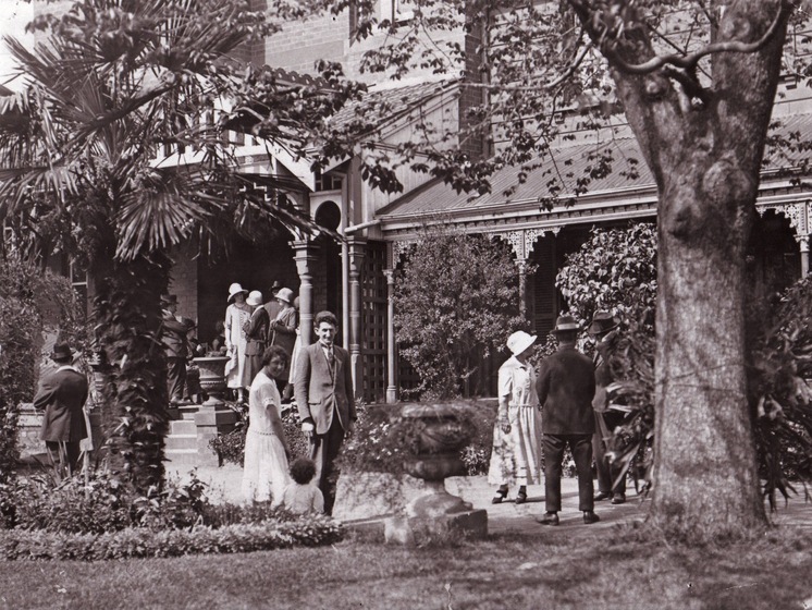 Black and white photograph of small groups of people conversing in the garden in front of a grand house.