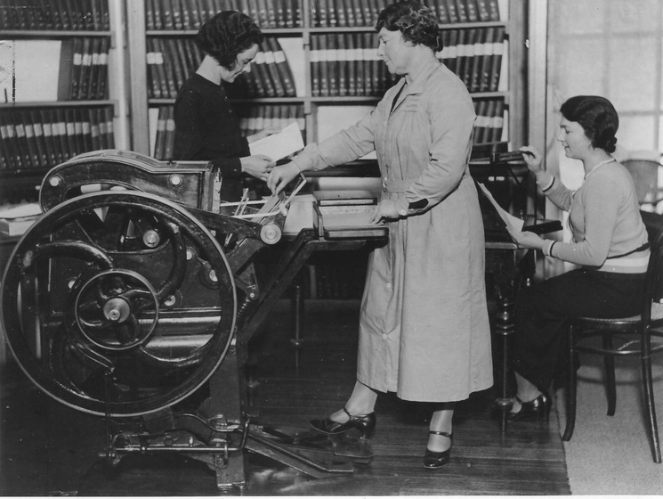 Black and white photograph of a woman standing with her foot on the pedal of a large machine while two other women work in the background in a library.