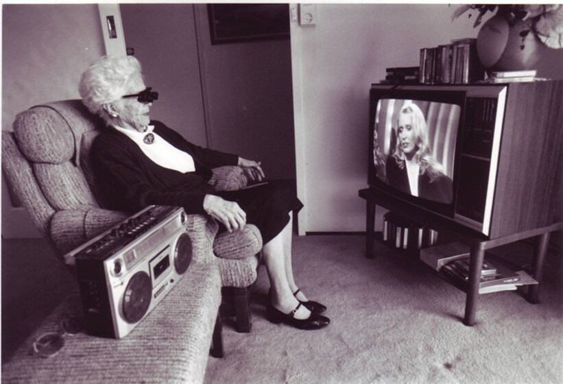 Black and white photograph of an elderly woman using assistive eyewear to watch television in a lounge room.