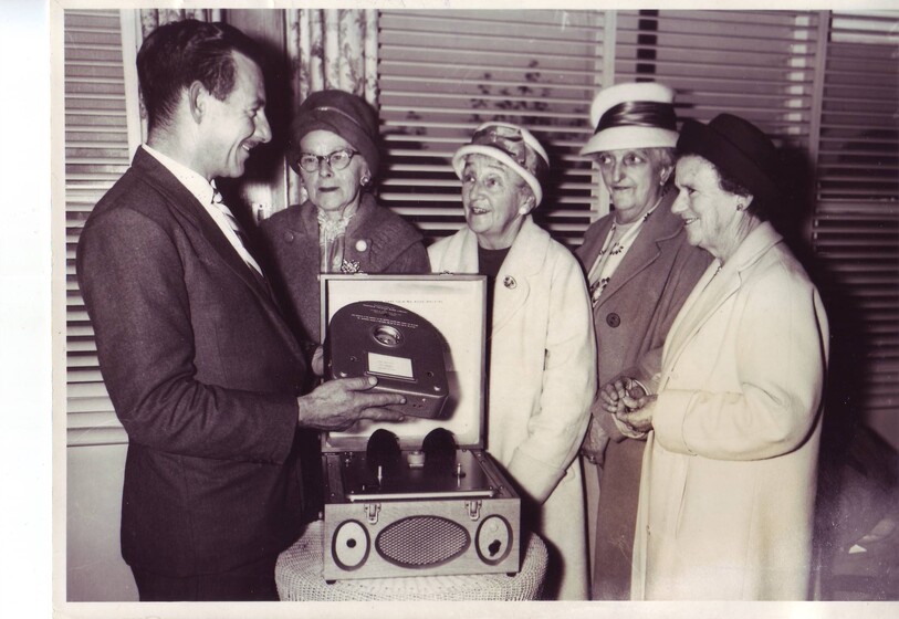 Black and white photograph man in a suit shows a piece of technology to four elderly women in hats.