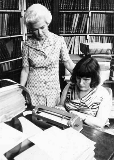 Black and white photograph of a woman sitting at a table using a braille typewriter while an older woman stands beside her in a library.