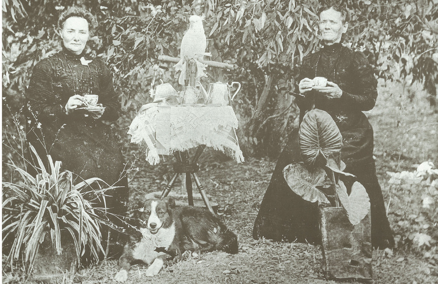 Black and white photograph of two women seated in a garden in black Victorian dresses, holding teacups, flanking a table with tea items, a cockatoo on a perch and a black and white dog.