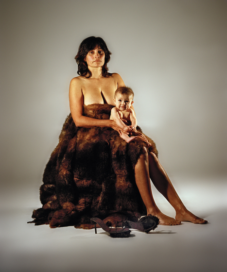 Colour photograph of a naked baby sitting on the lap of a naked woman draped in a fur cloak, a pair of stiletto shoes on the floor in front of her.