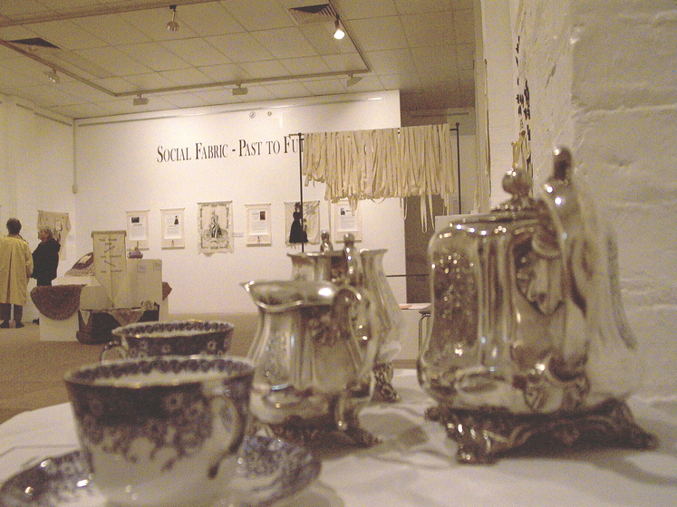 Colour photograph of gallery interior with silver tea service and tea cups in foreground and art works and patrons in background.