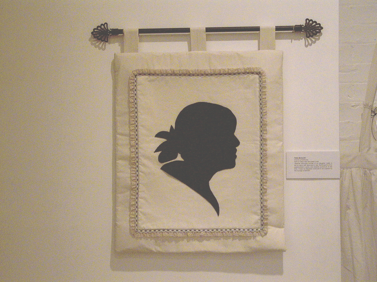 Small panel of white fabric framed with lace, hanging from a curtain rod, featuring the silhouette of a woman's profile.