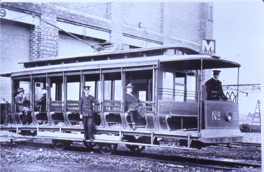 Black and white photograph of early open-sided electric tram outside depot with five men on board, including driver.