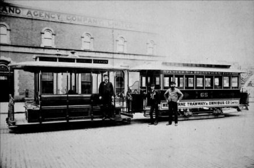 Black and white photograph of three men standing in front of two trams parked outside a Victorian building.