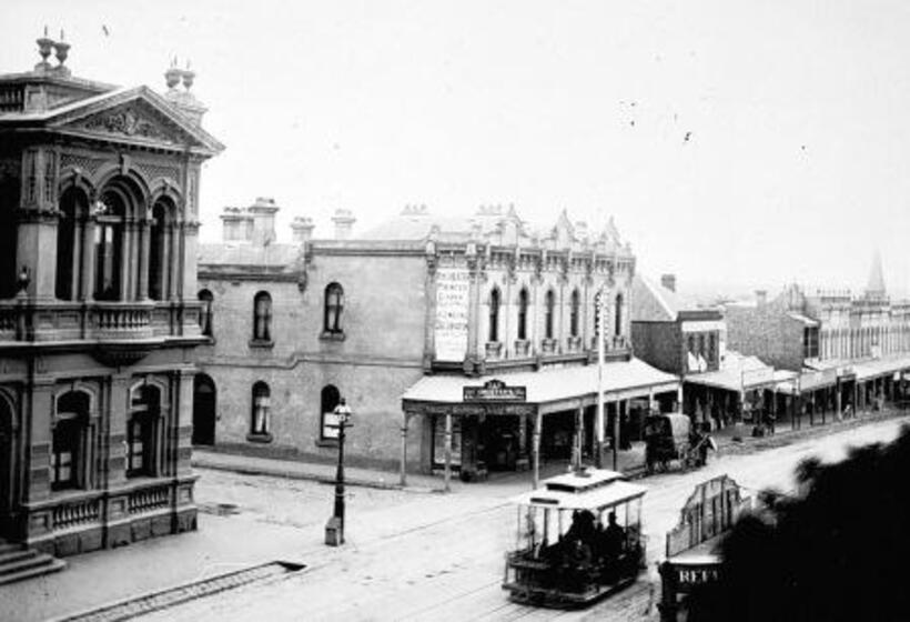 Black and white photograph of an open cable car travelling along a street of Victorian era shop fronts.