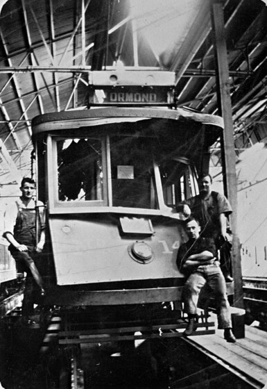 Black and white photograph of the front of a badly damaged tram in a workshop with three tradesmen posed on it.