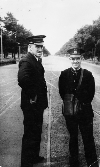 Black and white photograph of two men in tram uniforms standing on tram lines in a tree-lined street.