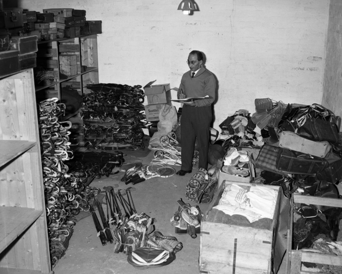 Black and white photograph of a man with an open folder and pen standing in a room surrounded by piles of sorted objects, including umbrellas, sports equipment, prams, suitcases, shopping bags and footwear.
