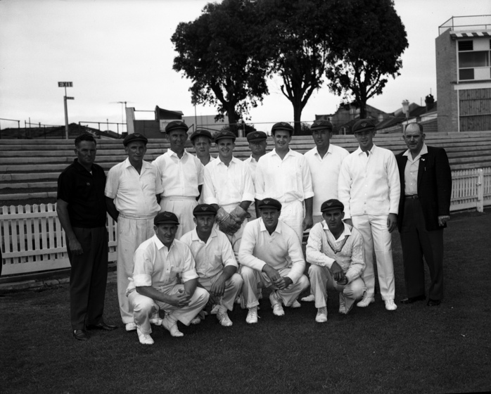 Black and white photograph of 12 men in cricket whites in two rows flanked by two men standing at the edge of a sports field with a white picket fence behind.