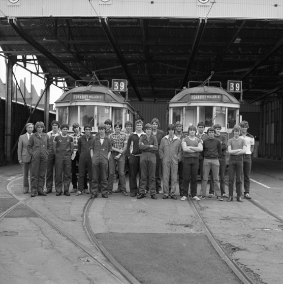 Black and white photograph of twenty young men and an older man in a suit standing in two rows in front of two trams in an open shed. Both trams labelled, 'Thornbury, Miller Street, 39'.