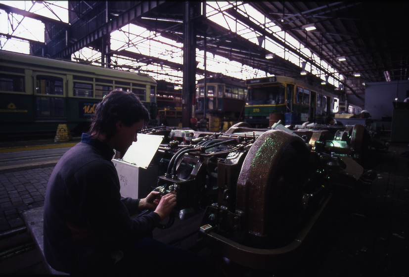 Colour photograph of a technician conducting electrical repairs to a tram undercarriage in a large workshop. Trams in the background.