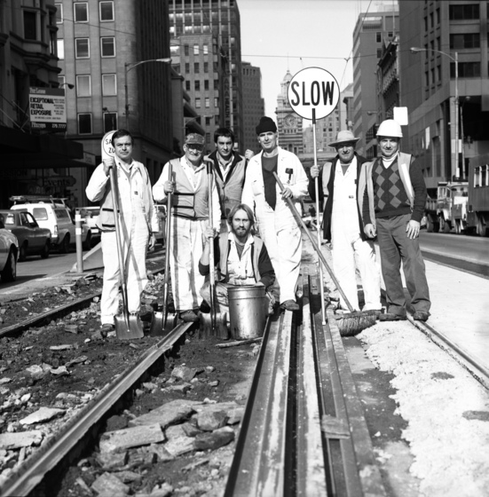 Black and white photograph of a row of six workmen standing and one squatting on tram tracks in the middle of a city road, holding brooms, shovels and a sigh saying, 'SLOW'.