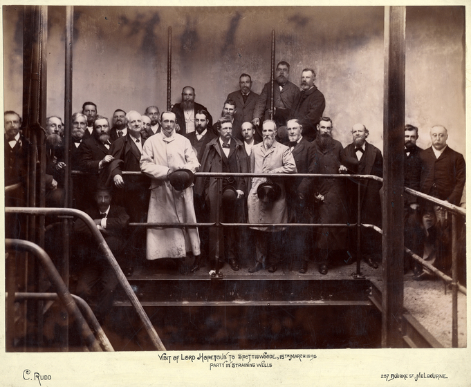 Black and white photograph of 24 men in Victorian dress standing at a metal railing.