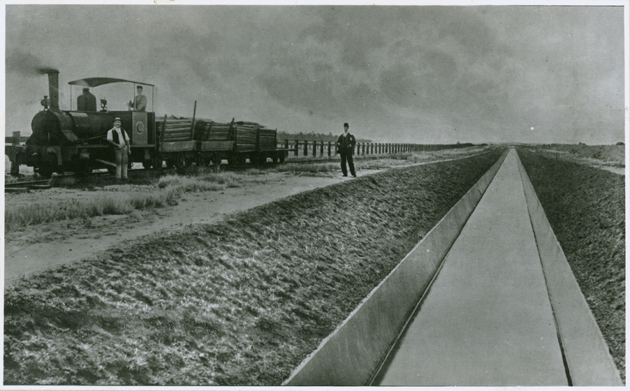 Black and white photograph of a small steam locomotive laden with timber beside a canal. Driver in locomotive. Man resting on locomotive. Man standing beside canal.