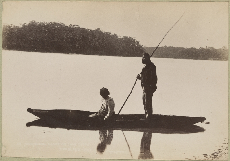 Black and white photograph of an Aboriginal man standing and an Aboriginal woman sitting in a canoe on a lake. Wooded shore in distance.