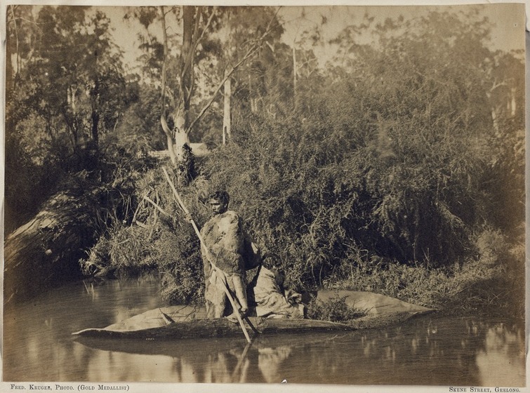 Black and white photograph of two Aboriginal people in fur cloaks in a bark canoe on a lake at the wooded shoreline. One standing, holding a pole. The other sitting.
