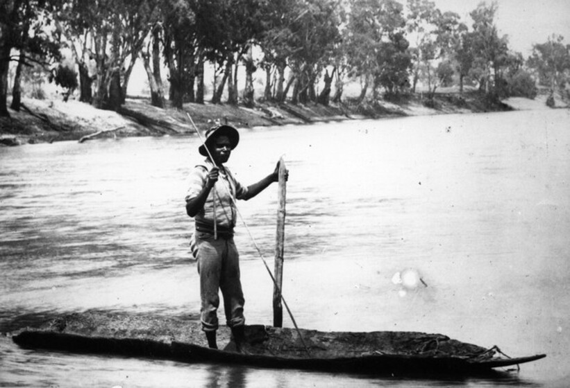 Black and white photograph of an Aboriginal man standing in a canoe on a lake, spear in on hand and pole in the other. Wooded shore in distance.