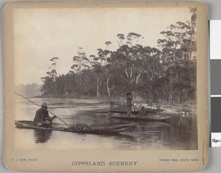 Black and white photograph of Aboriginal people in three canoes at the wooded shore of a lake.