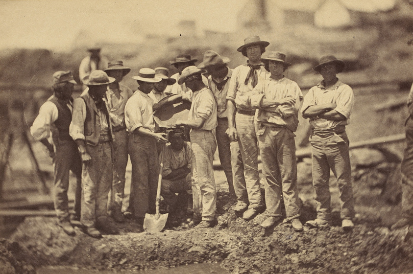 Group of miners standing on the goldfields panning for gold. Some of the men hold shovels, while others have pans.