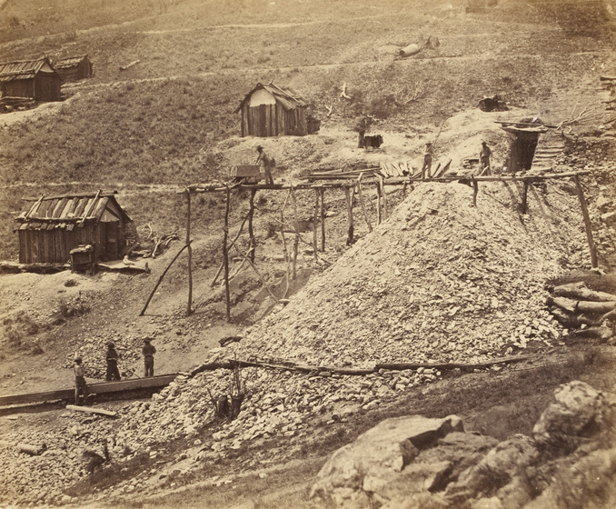 Sepia photograph of goldfields with wooden scaffolding on a tailings heap and bark huts.
