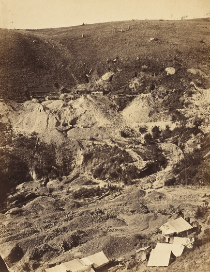 Sepia photograph of goldfield on bare hillside with tents and tailing heaps.