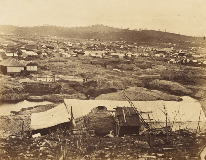 Sepia photograph of a goldfield with bare hills in the distance, tents, huts and tailings heaps in the middle ground and long white tent in the foreground.