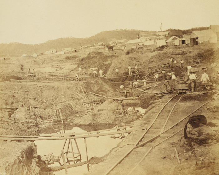 Sepia photograph of a goldfield with hills in the distance, huts on the rise in the middle distance and miners conducting extensive workings in the foreground, including wooden rails for hand wagons and pools of muddy water.