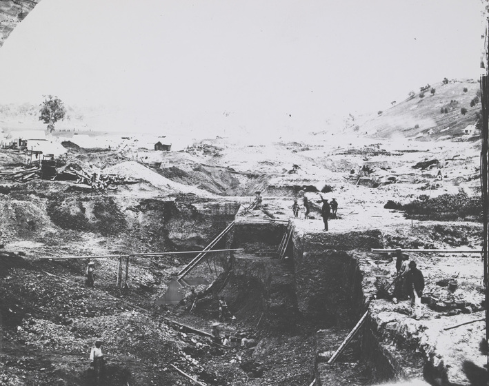 Black and white photograph of goldfield with miners looking into a deep hole in the workings.