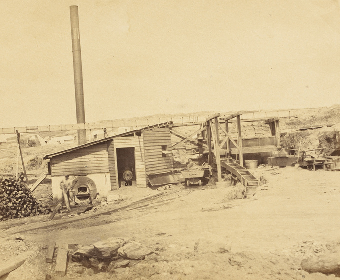 Sepia photograph of small weatherboard hut with furnace, tall metal chimney and sluice. A workman stands beside the furnace.