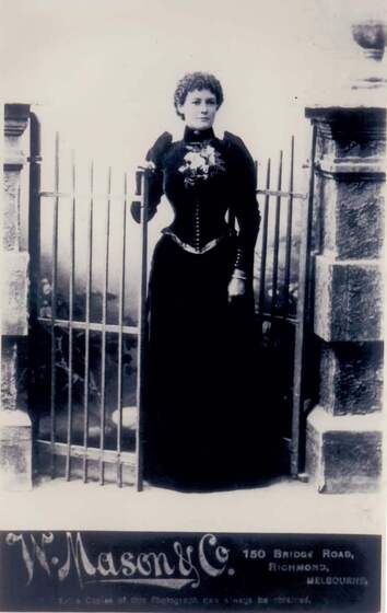 Black and white full length photo portrait of a young woman in a long dark Victorian gown.